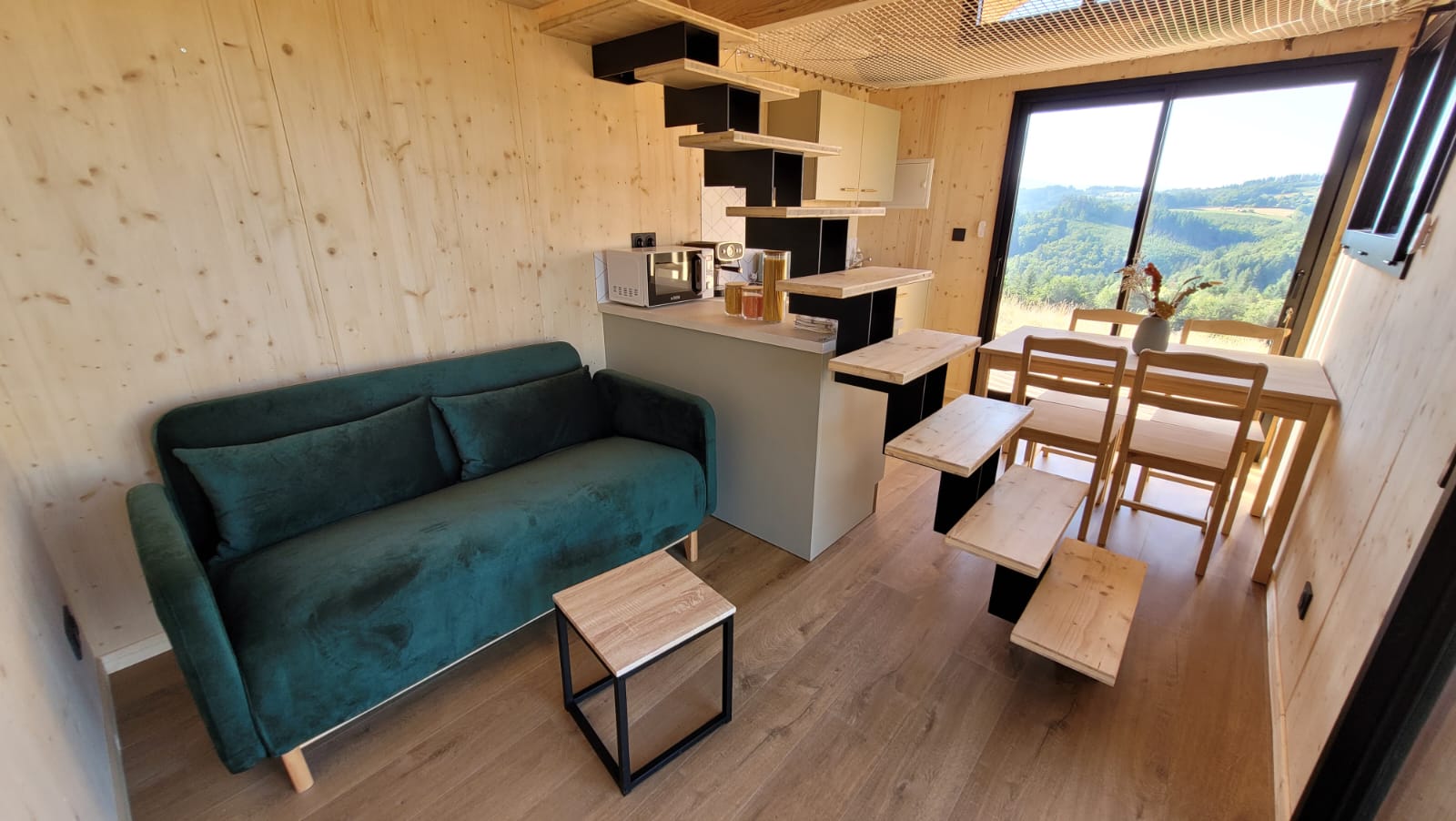 Salon avec canapé velours vert et cuisine ouverte, baie vitrée sur les collines